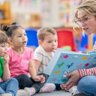 A preschool teacher sits on the floor of her classroom with a small group of students as she reads them a book. The children are each dressed casually and are focused on the story.