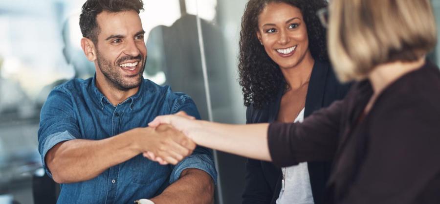 Three professionals sit together in a meeting, smiling as two people shake hands across a table in an office setting.