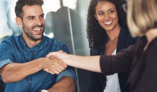 Three professionals sit together in a meeting, smiling as two people shake hands across a table in an office setting.