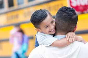 Young child holding father's neck while being carried to school bus